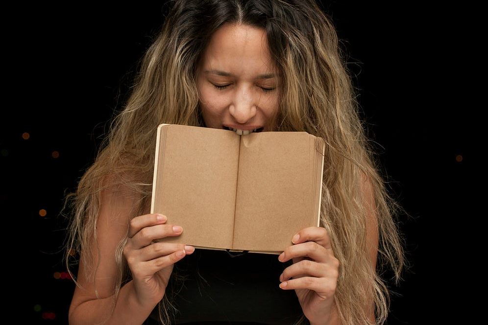 A woman playfully bites an open blank book, expressing a whimsical moment against a dark background.